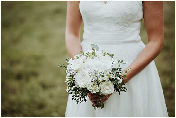 A close-up portrait of a bride from the torso down, holding a round bridal bouquet composed of ivory and white roses, white wax flower, small white spray roses, and trailing eucalyptus and olive-toned greenery. The bride wears a white strapless lace bodice gown with a flowy skirt. The bouquet is held with both hands at waist level against a softly blurred green outdoor background. The composition is a detail/portrait shot with a shallow depth of field that keeps the bouquet and dress as the focal point.