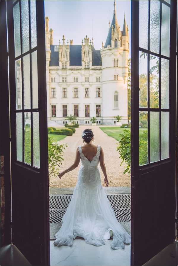 A bridal portrait taken from behind, framed by a set of open dark wooden double doors with glass panels. The bride stands on a threshold looking out toward a large French Gothic-style chateau with pointed turrets and ornate white stone facade, set along a gravel driveway lined with manicured hedges. She wears a fitted white lace gown with a low V-back, thin straps, and a trailing floor-length train, with her dark hair pinned up in an updo. The composition is a full-length portrait shot from inside the building, using the doorframe as a natural frame to draw the eye through to the chateau beyond. Potential venue feature image.
