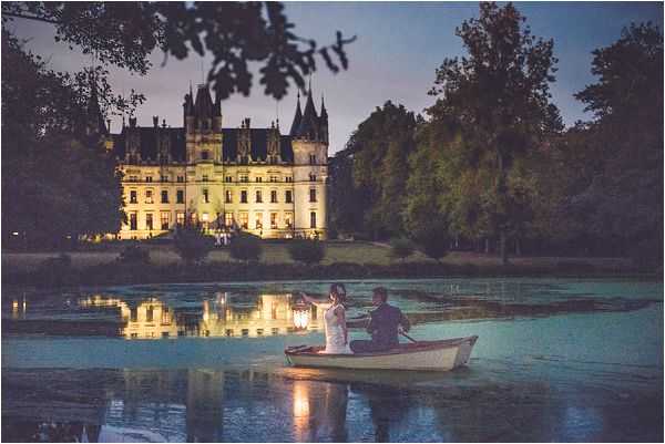 A couple sits in a small white rowing boat on a calm lake at dusk, with the bride holding a lit lantern that reflects on the water's surface. The groom, dressed in dark clothing, holds the oars while the bride wears a white dress. In the background, a large illuminated château with Gothic-style turrets and multiple lit windows is reflected in the lake, creating a mirror effect on the still water. The wide-angle shot captures the full scene from a low perspective, with tree branches framing the top of the image and a deep blue twilight sky overhead. Potential venue feature image.