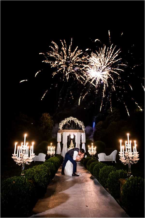 A couple shares a dip kiss on a paved garden pathway at night while a fireworks display bursts overhead in gold and white. The groom, dressed in a dark navy suit, dips the bride, who is wearing a white ballgown. The pathway is flanked by trimmed boxwood hedges and large crystal candelabras holding multiple lit white candles, casting warm light along the aisle. In the background, a white pavilion or gazebo decorated with white floral arrangements and draping curtains anchors the scene, with two white lounge sofas positioned on either side. The decor palette is classic and formal, combining white florals, crystal candelabras, and candlelight. Wide portrait shot with dramatic nighttime lighting from the fireworks and candles combined.