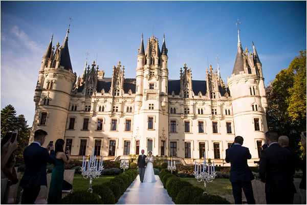 An outdoor wedding ceremony taking place directly in front of a large French Gothic-style château with white stone façade, pointed turrets, and ornate architectural detailing. The couple stands at the far end of a white aisle runner, flanked by tall silver candelabras holding blue and white floral arrangements, with neatly trimmed hedgerows lining the path. The bride wears a full-length white gown and the groom is in a dark suit; approximately 10–15 guests are visible on either side of the aisle, some holding cameras or phones. The shot is a wide-angle perspective taken from behind the guests, emphasizing the symmetry of the aisle and the full scale of the château as a backdrop. Potential venue feature image.