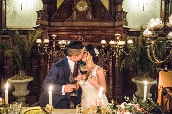 A bride and groom share a kiss at their sweetheart table during a wedding reception inside a grand historic interior space featuring ornate dark wood carved paneling, a large decorative mantelpiece with an antique clock, and tall candelabras with cream-colored shades. The groom wears a navy suit with a burgundy tie, and the bride wears a lace sleeveless gown with a white hair accessory; both hold glasses of red wine. The table is lit by tall white taper candles and decorated with a low floral arrangement of blush and peach roses with trailing greenery and eucalyptus, alongside gold tableware. Large stone urns with tropical palm fronds flank the couple on either side, contributing to a dark, moody, classic aesthetic. This is a medium portrait-style shot taken at table level.