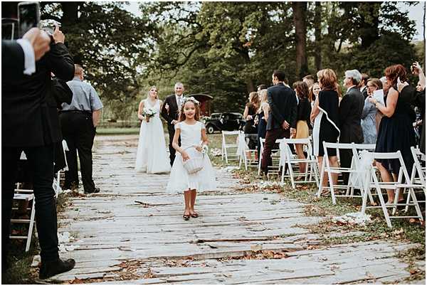 An outdoor wedding ceremony processional captured in a wide shot, showing a flower girl in a white tea-length dress walking down a wooden plank aisle ahead of the bride and her escort. The flower girl carries a small white basket and wears a white hair accessory. Behind her, the bride wears a long white gown and carries a small bouquet, accompanied by an older gentleman in a dark suit. Guests, approximately 30-40 people, are seated and standing on either side of the aisle in white folding chairs, with many turning to watch and photograph the procession. A photographer on the left side of the frame captures the moment. The setting is an outdoor venue with a tree-lined backdrop and a vintage dark-colored car visible in the background, suggesting a French chateau or estate grounds. The overall styling is classic and understated, with a natural autumnal feel indicated by scattered leaves on the aisle.
