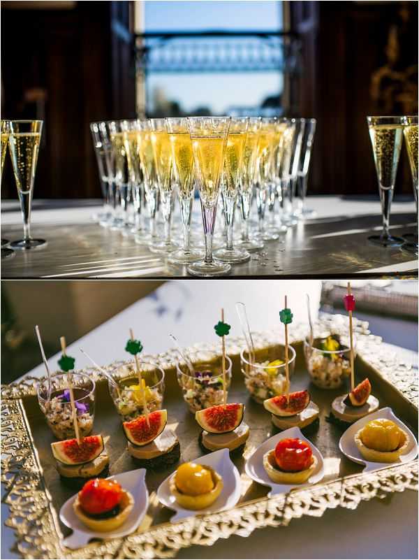 A two-panel detail shot of cocktail hour food and drink at a wedding reception. The top image shows rows of champagne flutes filled with golden sparkling wine arranged on a polished silver tray, backlit by a large window with architectural ironwork visible in the background. The bottom image shows a gold ornate tray presenting an assortment of canapés, including fresh fig slices on dark bread, small tartlets topped with glazed fruit in yellow and red, and individual clear plastic cups filled with a grain or rice-based salad garnished with edible purple flowers and colorful decorative picks in green, yellow, and pink.