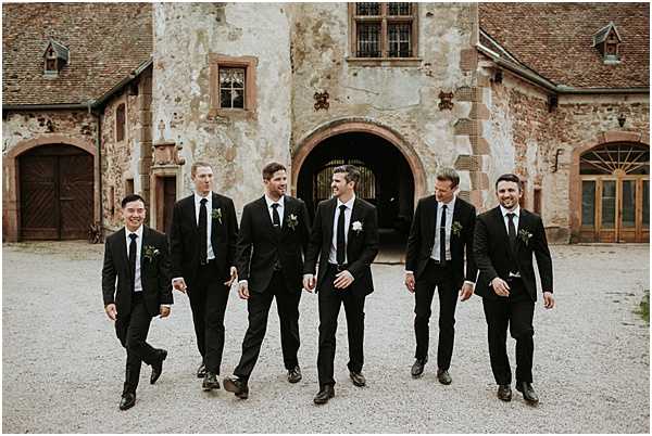 A groom and five groomsmen walk together across a gravel courtyard in front of a historic stone and brick château entrance featuring a large arched gateway with wrought iron gates. All six men wear matching black suits with black ties and white dress shirts, each with a small boutonniere featuring greenery and a white floral element. The groom appears to be the tallest figure in the center-left of the group, and the men are laughing and walking casually toward the camera in a wide, candid portrait shot. The styling is classic and coordinated with a monochromatic black palette. Potential venue feature image.
