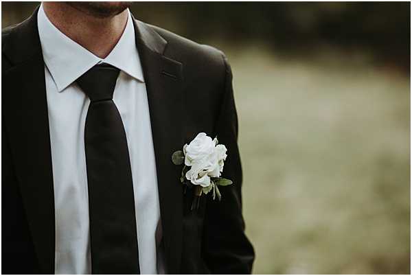 Close-up detail shot of a groom's lapel boutonniere, featuring two white roses with small green eucalyptus leaves, pinned to a dark charcoal suit jacket. The groom is wearing a white dress shirt with a black tie, creating a high-contrast, classic monochromatic look. The background is blurred into a soft neutral tone, keeping full focus on the boutonniere and suit styling. The overall aesthetic is clean and modern with a minimal floral approach.