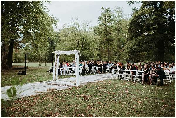 An outdoor wedding ceremony is underway on a grassy estate or park setting, with approximately 60-80 guests seated in white folding chairs arranged in rows on either side of a central aisle. The aisle is lined with a light-colored, possibly cream or off-white runner, and leads toward a simple white wooden arch decorated with white fabric draping and small floral accents. Guests are dressed in a mix of casual and semi-formal attire in varied colors. The ceremony appears to be at the moment just before or during the officiant's proceedings, as no couple is yet visible at the arch. Wide shot taken from behind the guests, capturing the full scene including the arch and surrounding grounds.