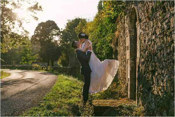 A couple portrait taken outdoors at golden hour, with the groom lifting the bride as they kiss beside a vine-covered stone archway along a rural road. The groom wears a dark navy or black suit, and the bride wears a white or ivory ballgown-style dress with a full skirt that flows outward from the lift. Warm late-afternoon backlight creates a strong sun flare effect and illuminates the scene with golden tones. The composition is a medium-wide portrait shot capturing both figures in full, with the stone arch and road providing depth and context in the frame.