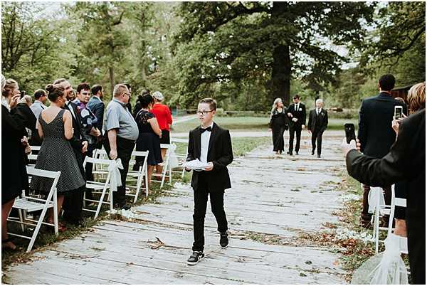 An outdoor wedding ceremony processional is underway along a wooden boardwalk aisle lined with white folding chairs and white ribbon accents. A young boy dressed in a black tuxedo with a black bow tie walks down the aisle carrying a white pillow or small tray, likely serving as a ring bearer. In the background, the wedding party — including several men in dark suits and what appears to be the bridal party — begins to make their way down the aisle. Guests standing on either side include a woman in a black and white polka-dot dress and others in casual to semi-formal attire, with one guest capturing the moment on a smartphone. The setting is a tree-lined outdoor venue with a rustic, natural feel, and the overall style appears relatively relaxed and informal. Wide shot taken from ground level along the aisle.