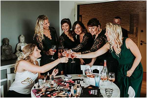 The bride and her bridal party are toasting with glasses of rosé champagne during the getting-ready phase of the wedding day. The bride is seated at a round table covered in makeup products and beauty tools, wearing a white tank top with 'Bride' written in gold lettering, while three bridesmaids in matching black lace long-sleeve dresses and one woman in a deep forest green dress stand around her raising their glasses. A bottle of rosé champagne is visible on the table alongside the spread of cosmetics. The setting appears to be a modern interior space with a stone accent wall and a decorative Buddha statue visible in the background. The shot is a candid medium-wide group portrait capturing a joyful, informal moment.