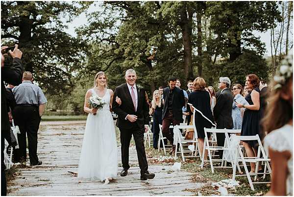 A bride is being walked down the aisle by an older man, likely her father, during an outdoor ceremony. The bride wears a flowing white A-line gown with lace detailing and carries a bouquet of white flowers with greenery. The aisle is paved with stone slabs lined with white folding chairs filled with approximately 30-40 guests, some of whom are standing and photographing the moment. The setting is a tree-lined outdoor ceremony space with a rustic, natural aesthetic, and scattered flower petals are visible on the ground along the aisle. Wide shot capturing the full procession from behind the guests in the foreground.