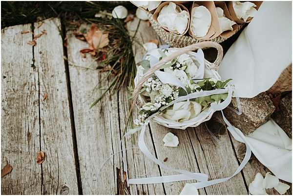 Close-up detail shot of flower girl baskets placed on weathered wooden decking, with scattered white rose petals around them. The small woven baskets are filled with white roses, baby's breath, and eucalyptus greenery, tied with white satin ribbons. Additional brown paper cones filled with white rose petals are visible in the upper portion of the frame, along with the edge of a white bridal gown. The styling is rustic and natural, with a white and green floral palette.
