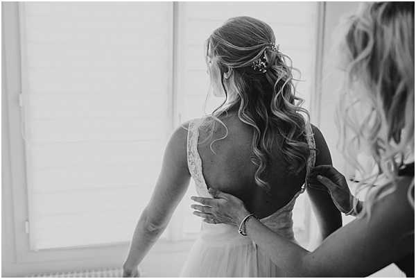 Black-and-white getting-ready shot of a bride being helped into her wedding gown by a bridesmaid or family member. The bride faces away from the camera, revealing a deep open back on her lace-trimmed dress, with soft light coming through shuttered windows behind her. Her hair is styled in loose, voluminous curls worn half-up, accessorized with a small decorative hair pin. The composition is a close-up portrait with moderate contrast, capturing the fastening detail at the back of the dress.