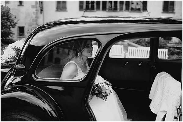 A black-and-white portrait of a bride seated inside a vintage classic car, likely a Citroën Traction Avant, decorated with flowers on the hood. The bride is smiling and looking forward, wearing a white lace or embroidered dress and holding a round bouquet with mixed blooms and foliage. A white embroidered or eyelet fabric — possibly part of her dress or a wrap — is visible draped over the car door. The shot is taken from outside the car at close range, framing the bride through the rear side window, with a stone building visible in the soft-focus background. The image has strong tonal contrast with deep blacks on the car body against the lighter tones of the bride and building.