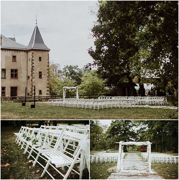 A three-image collage showing an outdoor wedding ceremony setup on the grounds of a French chateau with a distinctive pointed stone tower. The ceremony area features rows of white folding chairs arranged in two sections along a stone-paved aisle, leading to a white wooden arch draped with sheer white fabric and accented with small floral arrangements. The top image is a wide shot showing the full ceremony layout against the chateau facade, while the bottom-left detail shot shows white folding chairs adorned with small greenery and white floral accents along the aisle. The bottom-right image provides a straight-on view down the stone aisle toward the arch, showing the full seating arrangement. The overall decor palette is white and green with a classic, understated style. No guests or wedding party are present, indicating these are pre-ceremony setup shots. Potential venue feature image.