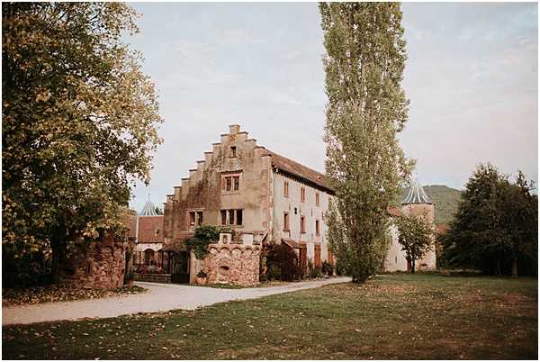 Wide shot of a historic French chateau or manor house featuring a stepped gable roofline, aged stone and rendered facade, and a round tower visible to the right. The grounds include a gravel driveway, a low stone wall with climbing foliage, and an open lawn in the foreground. No people are visible in this image. The autumn season is suggested by the yellowing leaves on the surrounding trees. Potential venue feature image.