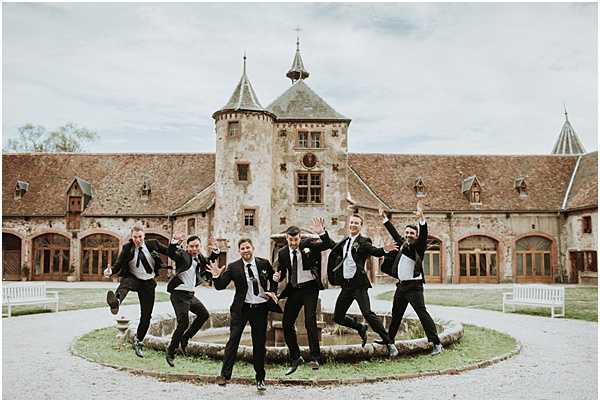 A group of seven men — the groom and his groomsmen — are caught mid-jump in a playful, energetic pose in the courtyard of a French chateau. All are dressed in black suits with white dress shirts and dark ties, with the groom distinguished by a floral boutonniere. The shot is taken in front of a historic stone chateau featuring a round tower with a pointed roof, terracotta-tiled roofing, arched doorways, and white wooden benches flanking the circular fountain they are jumping around. The wide shot captures the full courtyard and building facade, emphasizing the classic French architectural setting. Potential venue feature image.