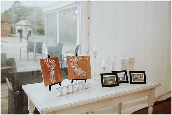 A close-up detail shot of a white wooden table styled as a welcome or memory display at an indoor wedding reception. The table holds two wooden string-art panels on easels labeled 'Alsace' and 'California,' referencing the couple's origins, alongside a white letter sign reading 'YOU & Me.' Two small framed photographs and a white candlestick holder with a lit pillar candle complete the arrangement. The decor palette is warm and rustic, mixing natural wood tones with white accents. Through the large window behind the table, an outdoor terrace with grey rattan chairs and string lights is visible in rainy conditions.