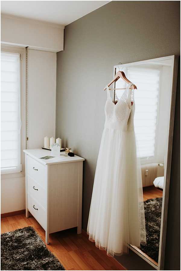 A getting-ready detail shot showing an ivory wedding dress hanging from a wooden hanger on a full-length floor mirror in a simply decorated bedroom. The dress features a lace bodice with thin straps and a flowing tulle A-line skirt. The room has grey-painted walls, warm wood flooring, a dark grey shag rug, and a white chest of drawers topped with white pillar candles and a few small beauty items. Natural light enters through shuttered windows. The composition is a medium wide shot capturing the full dress alongside the mirror's reflection of the room.