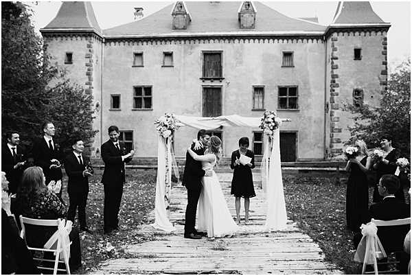 A black-and-white image of an outdoor wedding ceremony taking place on a stone or wooden pathway in front of a large historic chateau with pointed turrets and a rendered facade. The couple shares their first kiss at the altar, which consists of two tall standing posts draped with flowing white fabric and topped with floral arrangements featuring light-toned blooms. An officiant stands to the right of the couple holding papers. Guests are seated in white folding chairs on either side of the aisle, while the bridal party — groomsmen in dark suits on the left and bridesmaids in dark knee-length dresses on the right — stand and applaud. The image is a wide shot with moderate contrast, capturing approximately 20–30 attendees. Potential venue feature image.