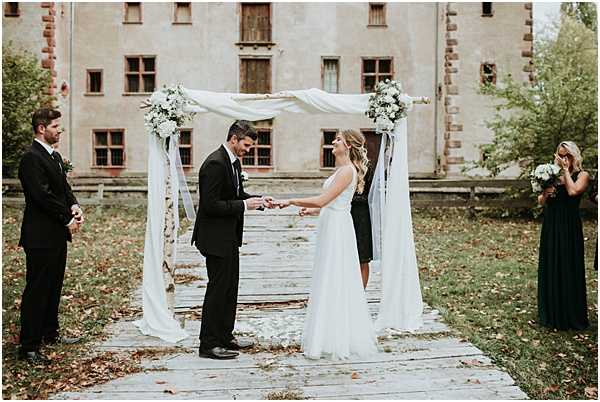 An outdoor wedding ceremony showing the couple exchanging rings beneath a square arch draped with flowing white fabric and decorated with white floral clusters, likely white roses and greenery, at the four corners. The groom wears a black tuxedo and the bride wears a simple white sleeveless A-line gown. The ceremony takes place on a rustic wooden platform with autumn leaves scattered around. A male officiant or groomsman stands to the left in a black suit, and a bridesmaid to the right in a floor-length dark forest green dress holds a white bouquet and appears emotional. The backdrop is a weathered, aged building with red-brick architectural details and worn facades, suggesting a historic chateau or manor. The overall styling is minimal and classic, with a black, white, and green color palette. Wide shot capturing all four figures and the full arch structure.