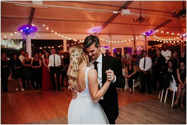 A couple shares their first dance on a wooden dance floor inside a white marquee tent, surrounded by a large group of seated and standing guests watching from the perimeter. The bride wears a white gown with an open back and has long wavy blonde hair adorned with a floral hair accessory, while the groom is dressed in a black suit with a white shirt and black tie. The tent is strung with warm-toned globe fairy lights along the ceiling, creating a golden ambient glow, with colored purple and blue uplighting visible in the background. The image is a medium wide shot taken from behind the bride, capturing the groom's face as he looks down at her.