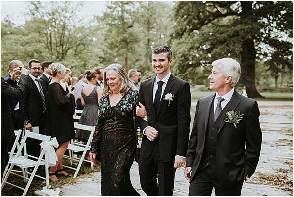 A groom in a black suit with a white boutonniere walks down the outdoor ceremony aisle flanked by two older adults, likely his parents — a woman in a long-sleeve black floral-embroidered dress and a man in a dark suit with a greenery boutonniere. White folding chairs line both sides of the aisle, with approximately 20–30 guests visible in the background dressed in dark formal attire. The ceremony takes place in a parklike outdoor setting with large mature trees. The shot is a medium wide candid portrait capturing the processional moment.