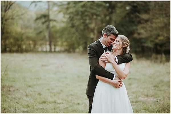 A couple portrait taken outdoors in an open field with a treeline in the background. The groom, wearing a dark olive/charcoal suit with a tie, stands behind the bride with his arms wrapped around her, both smiling and leaning toward each other. The bride wears a sleeveless white A-line gown with a fitted bodice and full skirt, with her hair styled in a loose updo. The shot is a medium portrait with a shallow depth of field, leaving the background softly blurred, and the overall color grading has a slightly muted, natural tone consistent with a boho or relaxed outdoor style.
