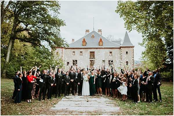 A large group portrait of approximately 50 guests gathered on the grounds of a French chateau, celebrating the bride and groom who stand at the center of the crowd. Guests are tossing white confetti into the air with arms raised, creating a festive celebratory moment. The bride wears a white ball gown and the bridesmaids are dressed in deep forest green gowns, while most male guests wear dark suits or tuxedos; a flower girl in a white dress is visible near the front. The chateau is a grey stone building with a blue-grey slate roof, turret, and multiple chimneys, set on a lawn flanked by mature trees, captured in a wide shot that centers the couple on a stone pathway leading to the building. Potential venue feature image.