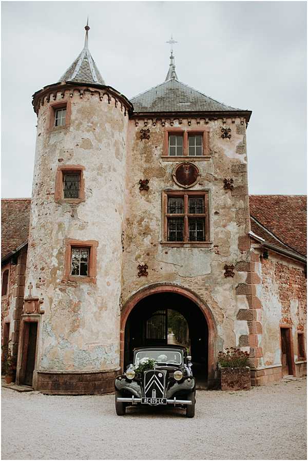 A vintage black Citroën Traction Avant, decorated with white floral arrangements on the hood, is parked in front of the arched gateway of a historic French chateau. The chateau features a round tower with a conical slate roof, weathered rendered walls with decorative brick detailing, and multi-pane leaded windows. The wide shot is taken from ground level, centering the vehicle beneath the arched entryway, with the full facade of the gatehouse filling the frame. No people are visible in the image. Potential venue feature image.