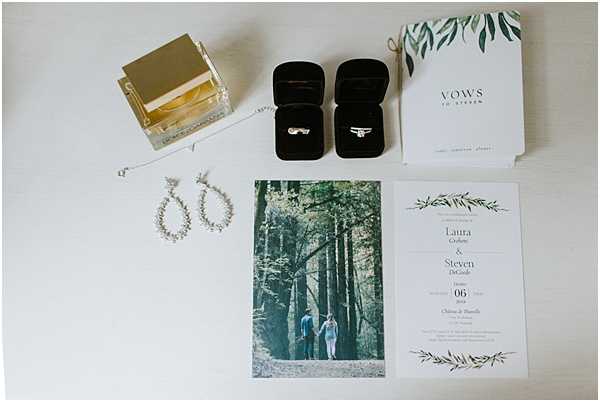 A flat lay detail shot of wedding stationery and accessories arranged on a white surface. The items include a wedding invitation suite with green botanical leaf illustrations for Laura and Steven's wedding at Château de Plumelec, a 'Vows to Steven' booklet with matching green leaf design, two black velvet ring boxes each containing a gold wedding band, a pair of delicate crystal drop earrings, a gold perfume bottle, and a photo card insert featuring the couple walking hand-in-hand through a wooded forest setting. The overall stationery palette is white and green with a botanical theme.