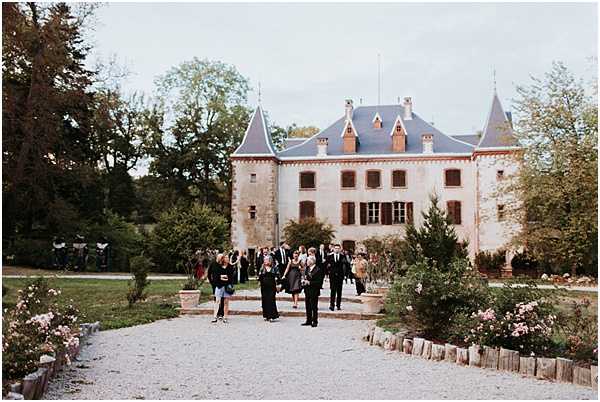 Wedding guests mill about on a gravel pathway in front of a French chateau during what appears to be a cocktail hour or post-ceremony gathering. The venue is a classic multi-story chateau with blue-grey slate roofs, corner turrets, and brown wooden shutters. Approximately 20–30 guests are visible, dressed in formal attire predominantly in dark and black tones. Rose bushes with pale pink blooms line both sides of the gravel path, edged with low wooden borders. The shot is a wide exterior view taken at dusk or in soft overcast light. Potential venue feature image.
