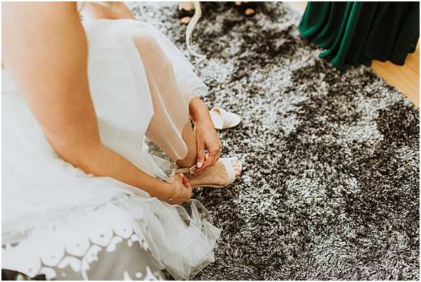 A close-up detail shot of a bride sitting on a dark grey shag rug fastening a nude or blush strappy heeled sandal onto her foot during the getting-ready portion of the day. She is wearing a white lace-hemmed gown, and a ring is visible on her finger. In the background, the lower portion of a person in an emerald green dress is partially visible, along with a second white shoe resting on the rug.
