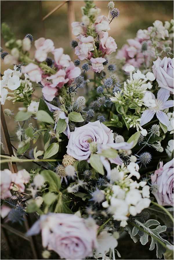 Close-up detail shot of a wedding floral arrangement featuring a loose, garden-style composition in a soft purple and mauve palette. The arrangement includes lavender and blush garden roses, pale pink snapdragons, blue-grey eryngium thistles, white clematis, and small white filler flowers, all interspersed with lush green foliage and dusty miller. The overall styling is romantic and organic with a wildflower aesthetic, shot with a shallow depth of field that keeps the central rose in focus while the surrounding blooms fall into a soft blur.
