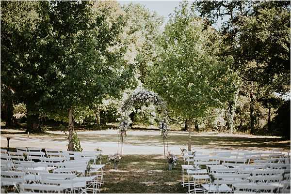 An outdoor ceremony setup photographed from the back of the aisle in a wide shot, showing rows of white folding chairs arranged on either side of a central aisle leading to a floral arch. The arch appears to be a circular or rounded metal structure adorned with white and soft lavender or mauve flowers and greenery, flanked by two smaller floral arrangements on stands at the entrance to the arch. The setting is a shaded garden or wooded clearing with large mature trees providing a natural canopy. The overall decor palette is soft white and muted purple-grey, consistent with a romantic garden-style ceremony aesthetic. No guests or wedding party are present, indicating this was taken before the ceremony began.