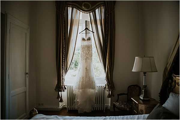 A getting-ready detail shot of an ivory lace wedding dress hanging from a curtain rod in front of a tall window inside what appears to be a chateau or manor house bedroom. The dress features a fitted silhouette with intricate lace detailing and a ruffled train. The room is decorated in a classic style with gold-toned draped curtains with tassels, a white cast-iron radiator beneath the window, a traditional bedside lamp, an ornate gilded bed frame partially visible on the right, and a floral-upholstered chair. The moody, low-contrast lighting creates deep shadows across the room, with natural backlight from the window illuminating the dress.