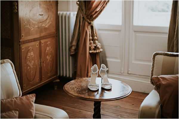 A detail shot of a pair of white bridal heels with ankle straps and delicate embellishment at the toe, placed on a small antique wooden side table with a twisted barley-twist pedestal base. The setting is an ornate interior room featuring a painted wooden armoire, a white cast-iron radiator, heavy gold-taupe drapery with tassel trim tied back at a tall French window, and cream upholstered armchairs with dusty rose cushions. The warm, slightly moody natural light from the window gives the image soft, golden tones. The composition is a medium shot emphasizing the bridal shoes as the focal point within the classic, period-furnished room.