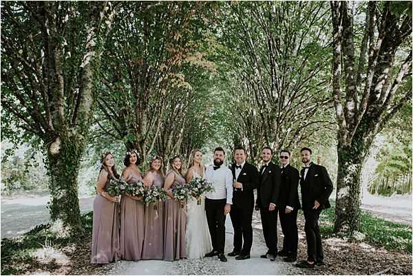 A bridal party group portrait taken outdoors along a tree-lined path, with tall trees forming a natural canopy overhead. The group consists of approximately nine people: the bride in a white lace gown, the groom in a white shirt and black trousers with a bow tie, four bridesmaids in mauve-taupe floor-length dresses carrying loose bouquets of dusty purple and greenery blooms, and three groomsmen in black suits. One bridesmaid wears a floral crown. The bouquets feature muted purple, mauve, and green tones in a loose, organic arrangement consistent with a boho styling aesthetic. The wide shot captures the full group posed casually and smiling, with dappled natural light filtering through the tree canopy.
