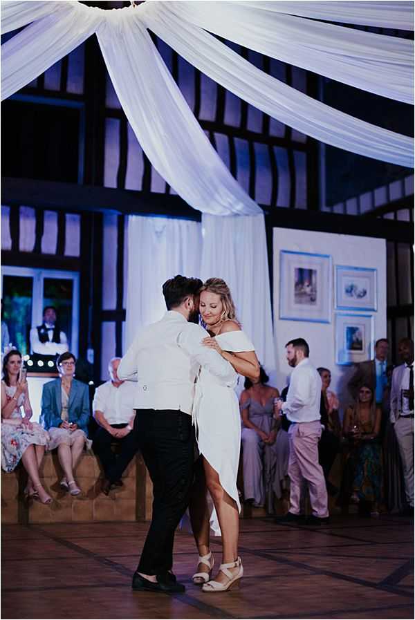 The couple shares their first dance in an indoor reception venue with exposed dark timber-frame beams and white fabric draping radiating from a central ceiling point, lit with blue ambient uplighting. The bride wears a short off-the-shoulder white dress with a high-low hemline and beige wedge sandals, while the groom is dressed in a white shirt and black trousers. Approximately 20-25 guests are seated and standing around the perimeter of the dance floor watching, dressed in smart-casual attire including a light blue blazer and a floral dress visible on the left. The image is a medium wide shot taken from a slightly low angle, with framed artwork visible on the back wall and a staff member in a black waistcoat standing in the background.