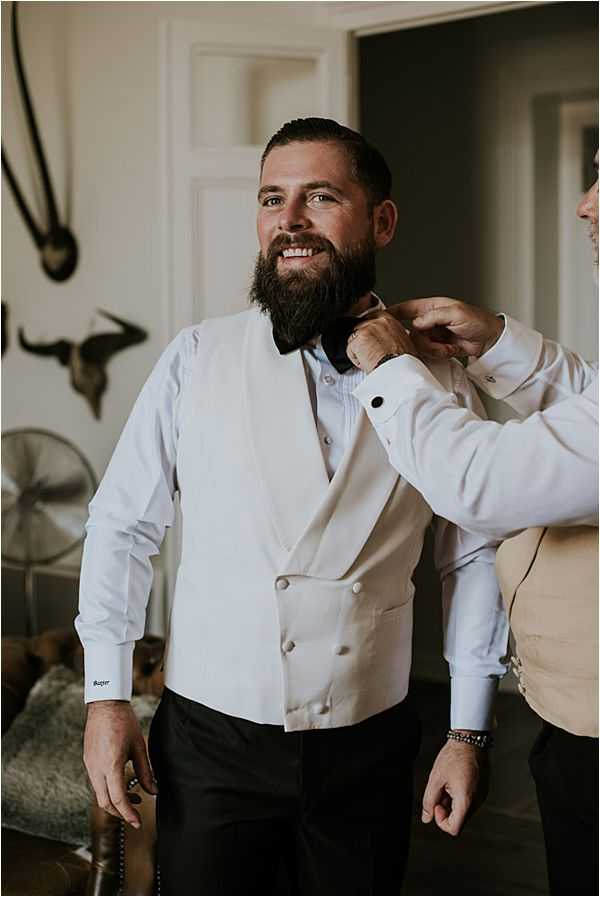 A getting-ready scene shot indoors, showing a bearded groom smiling as another man adjusts his dark bow tie. The groom is wearing a white double-breasted waistcoat over a white dress shirt with cufflinks, paired with black trousers. The assistant partially visible on the right is wearing a tan or champagne waistcoat. The room has a classic interior with white panelled walls, and animal skull wall decorations visible in the background, suggesting a chateau or manor house setting. The image is a mid-length portrait shot with warm, natural window light.