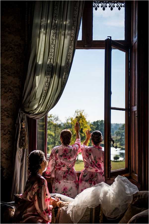 A getting-ready scene captured indoors at what appears to be a chateau, showing three women in matching pink floral satin robes gathered at a large, ornate double-casement window with dark wood frames and decorative ironwork detailing. Two of the women stand at the open window looking out over the estate grounds, with one raising her hand, while a third sits in the foreground. A white wedding dress is partially visible in the lower right corner of the frame. The room features patterned wallpaper, a heavy sage-green curtain with tassel trim, and warm natural light streaming through the window. The composition is a medium portrait shot taken from behind the subjects, with strong backlighting creating a silhouette effect against the bright exterior view.