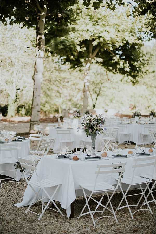 An outdoor wedding reception setup photographed in a wide portrait-style shot, showing multiple round tables dressed in white linen tablecloths arranged on a gravel surface beneath mature trees. White metal folding bistro chairs surround each table, and the nearest table features a centerpiece of blush pink, lavender, and purple blooms in a dark urn-style vase, with slate-colored charger plates and small orange accent pieces at each place setting. Globe string lights are strung between the trees overhead, and additional florals are visible on tables in the background, creating a cohesive soft, garden-party aesthetic with a white and muted pastel palette.