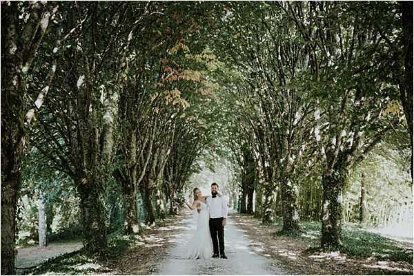 A couple portrait taken outdoors along a tree-lined driveway or allée, with tall trees forming a natural canopy overhead. The bride wears a white fitted gown with a train and the groom is dressed in a black tuxedo with a white shirt and bow tie; they stand holding hands facing the camera. The shot is a wide portrait with the couple positioned centrally at mid-distance, emphasizing the symmetrical row of trees stretching into the background. The overall styling is classic and the setting suggests a French chateau or country estate entrance.