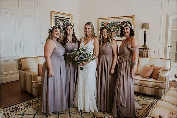 A group portrait of a bride and four bridesmaids posing together indoors in what appears to be a chateau salon or sitting room, with white paneled walls, framed artwork, and a sofa with cushions visible in the background. The bride wears a white lace gown with a V-neckline and holds a loose bouquet of white, blush, and soft blue flowers with greenery. The bridesmaids wear mismatched floor-length dresses in muted mauve and taupe tones, and three of the four wear floral crowns with small white and green blooms in their hair. The overall styling leans bohemian with the mix-and-match dress palette, flower crowns, and loosely arranged bouquet.