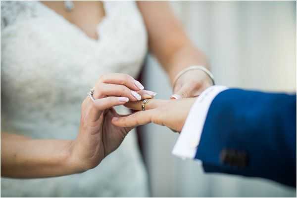 Close-up detail shot of a ring exchange during a wedding ceremony, showing the bride sliding a gold band onto the groom's finger. The bride is wearing a white lace dress and a delicate bracelet, with manicured nails in a pale pink polish, while the groom is dressed in a navy blue suit with a white dress shirt. The background is softly blurred, with neutral tones suggesting an outdoor or semi-outdoor setting.