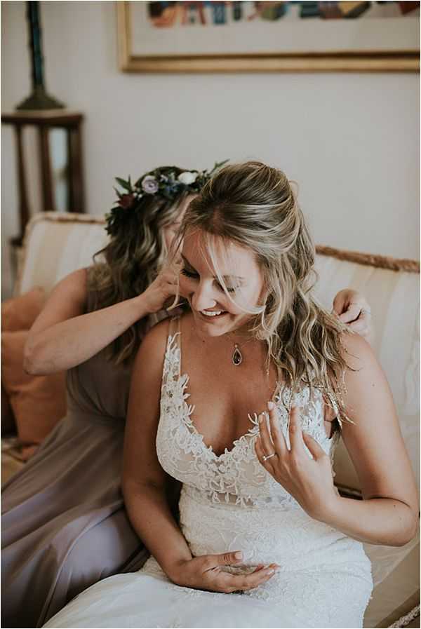 A getting-ready moment captured indoors, showing the bride seated on a cream upholstered chair or small sofa while a bridesmaid or friend helps adjust her hair from behind. The bride wears a white lace wedding dress with a deep V-neckline and floral appliqué detailing, paired with a teardrop dark gemstone pendant necklace. Her wavy blonde hair is worn half-up, half-down. The person assisting her wears a taupe or pale grey satin dress and a boho-style floral crown made of dark greenery, small white blooms, and muted purple flowers. The bride is smiling and laughing during the moment. A framed colorful artwork and a wooden lamp are visible in the background, suggesting a room in a house or villa. The styling leans toward a relaxed boho aesthetic. Medium close-up portrait shot.