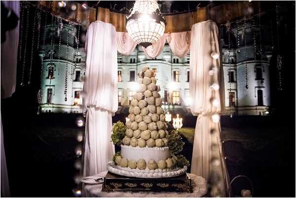 A traditional French croquembouche wedding cake is displayed outdoors at night, built as a tall tower of cream puffs on a decorative gold-trimmed stand, adorned with green hydrangea clusters at its base. The dessert table is framed by white draped fabric columns hung with crystal bead strands and fairy lights, with a crystal chandelier suspended above. In the background, a large illuminated chateau is lit up in blue-green tones, clearly visible against the dark night sky. The wide shot composition places the croquembouche as the central focal point with the chateau serving as a dramatic backdrop. Potential venue feature image.