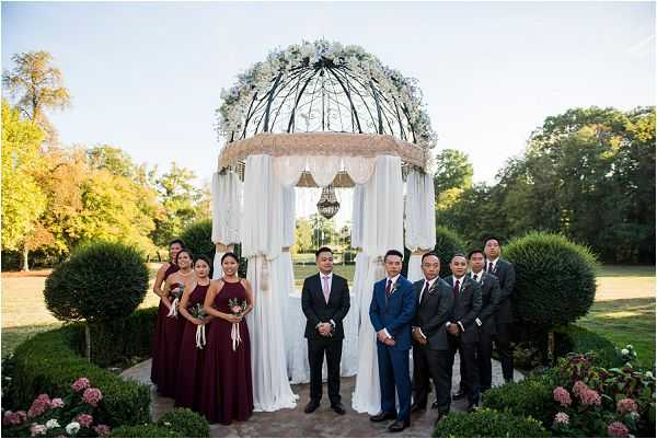 A bridal party portrait taken outdoors in a formal garden setting, with the group posed in front of an ornate wrought-iron gazebo ceremony structure. The gazebo is decorated with white draping, hanging crystal or lantern accents, and a crown of white floral arrangements at the top. The groom stands at center in a black suit with a light pink tie, flanked by groomsmen in navy blue suits with burgundy ties and pocket squares. Four bridesmaids stand to the left wearing floor-length burgundy halter-neck gowns and carrying small bouquets. Additional groomsmen and attendants flank both sides, totaling approximately 10-12 people in the wedding party. The wide-shot composition captures the full gazebo structure and the surrounding manicured garden with trimmed topiary shrubs and low flowering plants in the foreground.