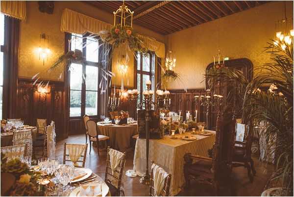 A wide shot of an indoor wedding reception room set up for a seated dinner, likely inside a historic chateau or manor house. The room features dark wood wall paneling, a coffered wooden ceiling, tall arched windows with gold-yellow drape valances, and warm amber lighting from wall sconces and chandeliers. Multiple round and rectangular tables are dressed in gold and ivory lace-style linens, set with crystal glassware, candles, and floral centerpieces incorporating greenery, palm fronds, and small white blooms. A tall ornate gold candelabra serves as a central decorative element, adorned with trailing greenery and white flowers. The decor palette is warm gold, ivory, and earthy green, with a classic, vintage-inspired styling theme. Approximately four to six tables are visible, each with chairs dressed in ivory or white chair covers. A large ornate mirror and carved wooden furniture pieces are visible in the background. Potential venue feature image.