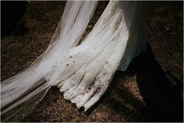 A close-up detail shot taken outdoors on grass, focusing on the train of a bride's ivory lace wedding dress and a long tulle veil spread across the ground. Small scattered red and dark confetti or flower petals are visible on the fabric and surrounding grass. The lace hem features intricate scalloped edging. The lower legs of the bride and a figure in dark trousers are partially visible at the top of the frame.